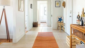 A hallway with a patterned rug, a lamp, framed pictures, and a wooden side table with bottles. - Cozy 2BR/2BA Apt on Broadway (Newport)