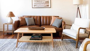 A living room with a brown leather sofa, a white armchair, a wooden coffee table, a floor lamp, and a framed picture on the wall. - Cozy 2BR/2BA Apt on Broadway (Newport)