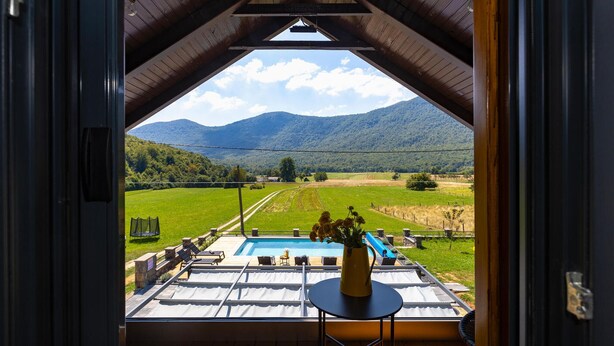 Vista de um quarto com teto de madeira e janela para uma piscina, campos gramados e montanhas.