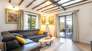 Uma sala de estar moderna com um sofá seccional, uma mesa de centro e um abajur. Há duas janelas com cortinas, um painel de vidro deslizante que dá para uma área externa e um quadro na parede.