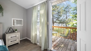 A bedroom with a wooden floor, a nightstand with drawers, a window with curtains, and a view of a wooden deck with outdoor furniture. - The Bohemian A frame. (Lampe)