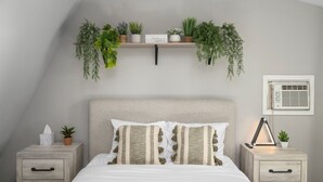 A bedroom with a bed, bedside tables, a shelf with plants, and an air conditioning unit. - The Bohemian A frame. (Lampe)