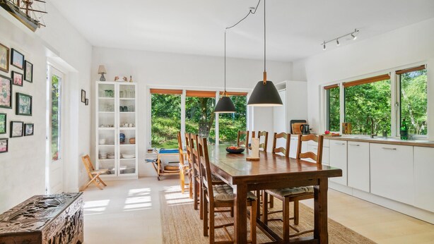 A spacious dining area with a long wooden table, surrounded by chairs, and a view of lush greenery through large windows.