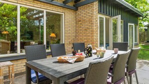 An outdoor patio with a wooden table and wicker chairs, a brick wall, and large windows.