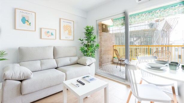 Una sala de estar moderna con un sofá, una mesa de centro y un comedor con sillas y una mesa. Hay obras de arte enmarcadas en la pared y un ventanal con vistas a un balcón.