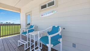 A covered outdoor seating area with white and teal chairs and a table, a sign above the chairs, and a view of a green field. - Bonnie Dune by Silver Sands Vacation Rentals (Port Aransas)