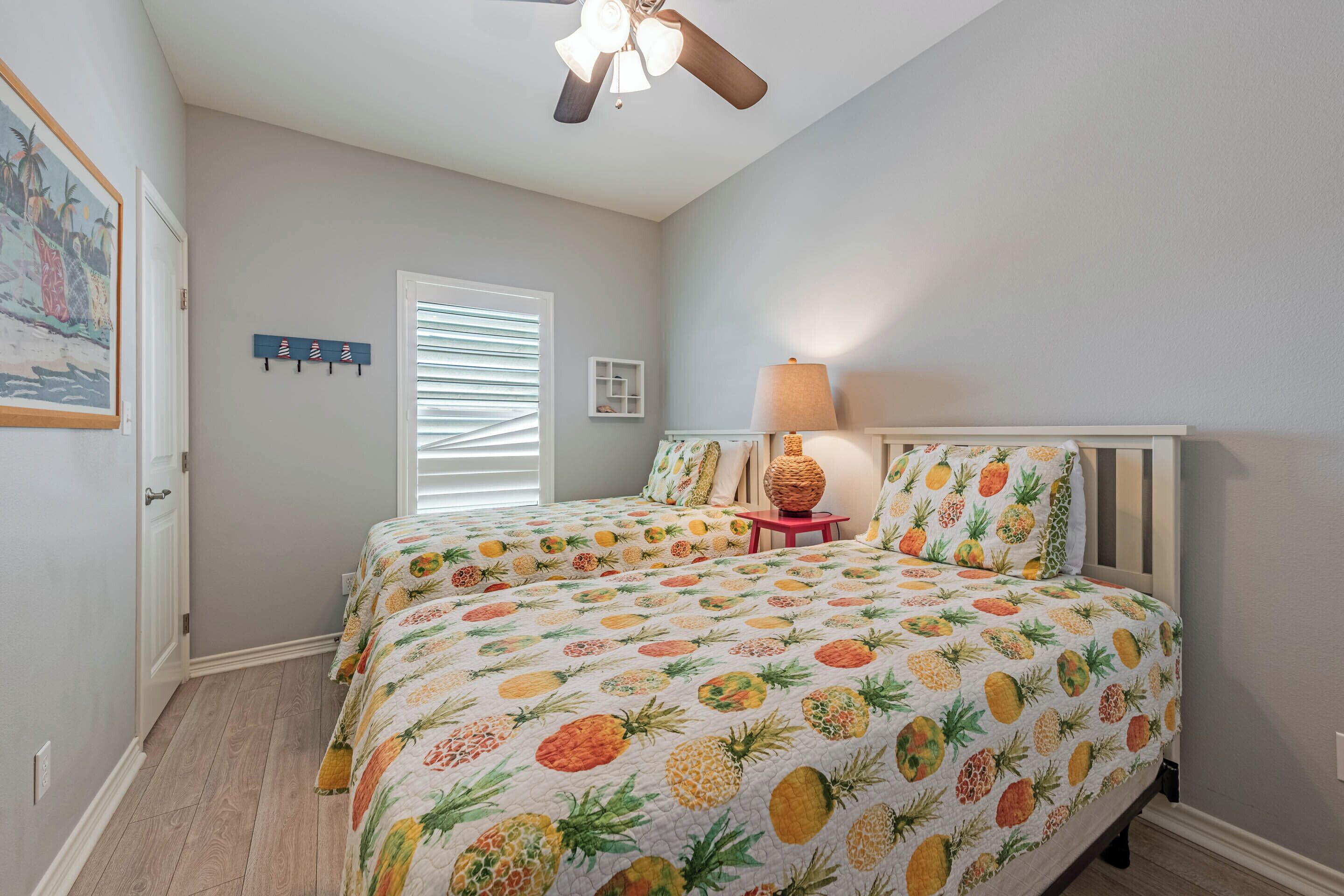 A bedroom with a bed featuring a pineapple-themed bedspread, a ceiling fan, a window with blinds, and a small table with a lamp.