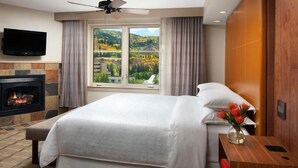 A hotel room with a bed, a fireplace, a television, and a window with a view of a forest and hills. - Sheraton Lakeside Terrace Villas at Mountain Vista (Avon)