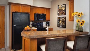 A kitchen with wooden cabinets, a black refrigerator, a microwave, a stove, and a granite countertop. There are two chairs and a vase with sunflowers on the counter. - Sheraton Lakeside Terrace Villas at Mountain Vista (Avon)