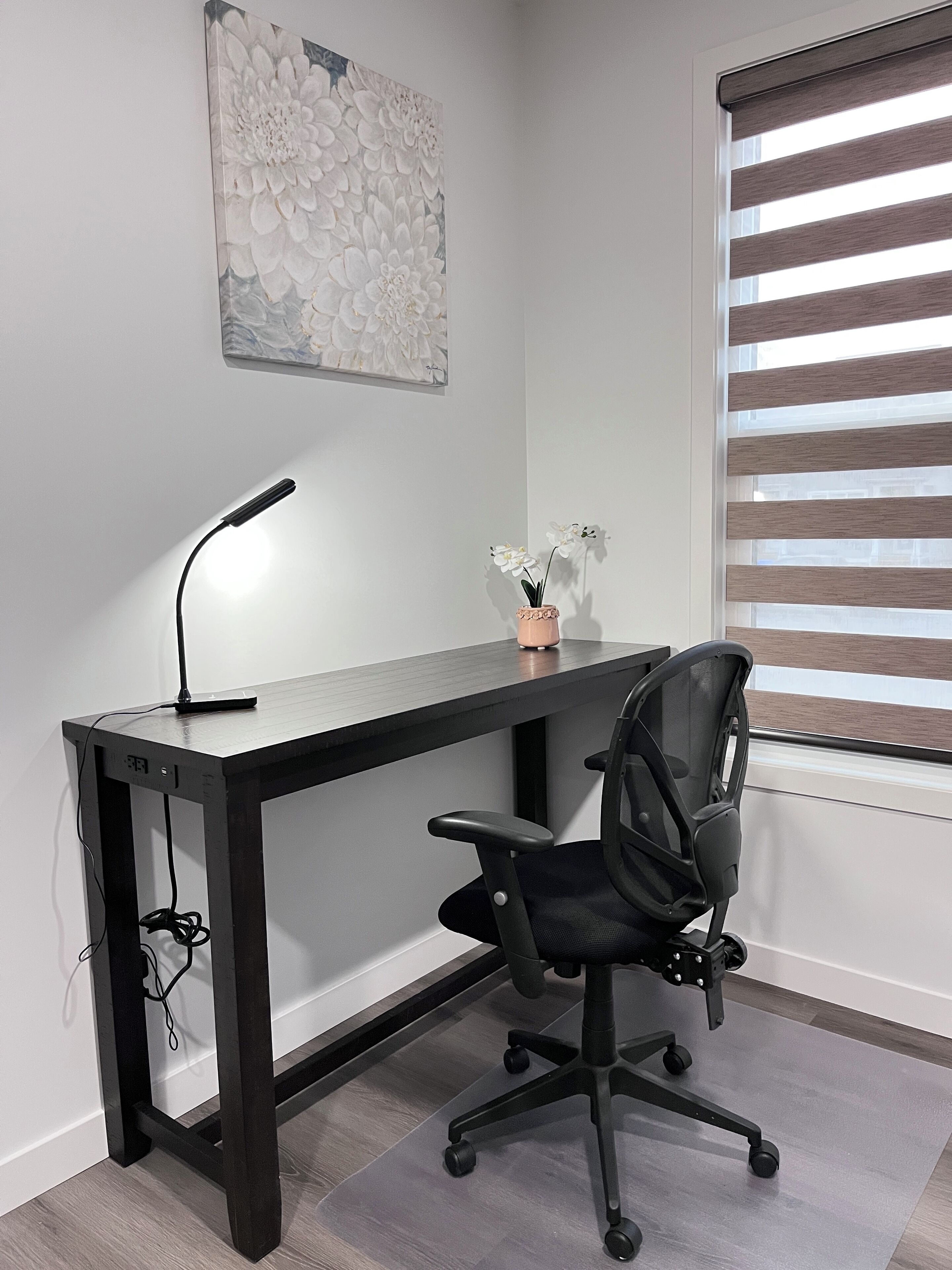 A modern office desk with a black chair, a desk lamp, and a vase with white flowers.