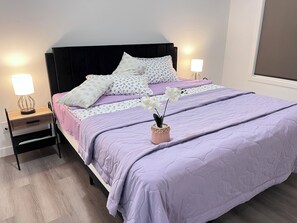 Uma cama bem arrumada com colcha roxa, duas luminárias de cabeceira, um criado-mudo com gaveta e uma orquídea em vaso sobre a cama.