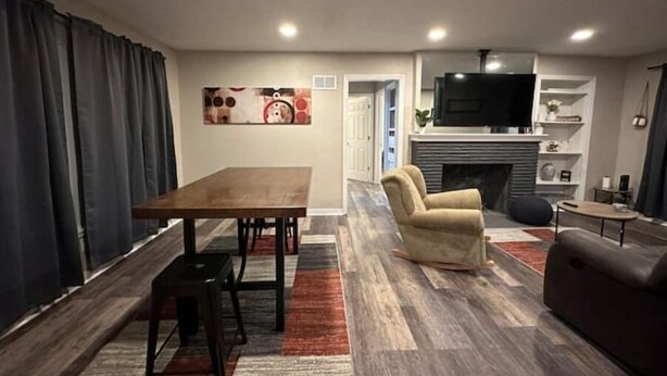 Een moderne woonkamer met een houten eettafel, een beige fauteuil en een tv boven een open haard.
