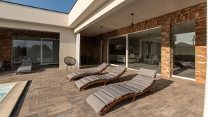 A modern outdoor lounge area with sun loungers, a pool, and a dining area visible through glass doors. - Cozy home in Seline with sauna (Seline)