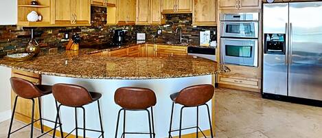 A modern kitchen with a central island, bar stools, wooden cabinets, stainless steel appliances, and a backsplash with a mosaic tile design.