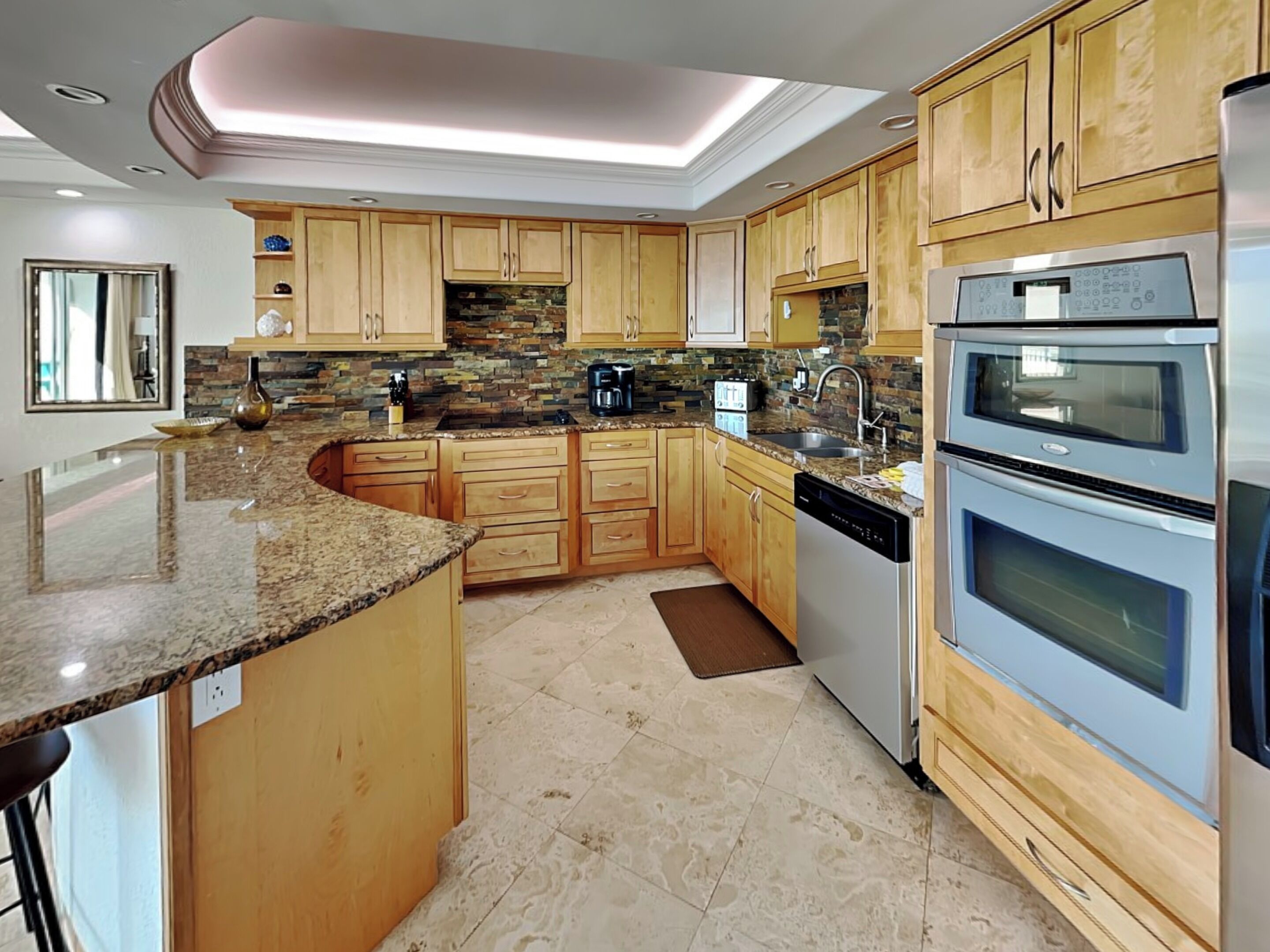 A modern kitchen with granite countertops, wooden cabinets, a built-in oven, and a stone backsplash.