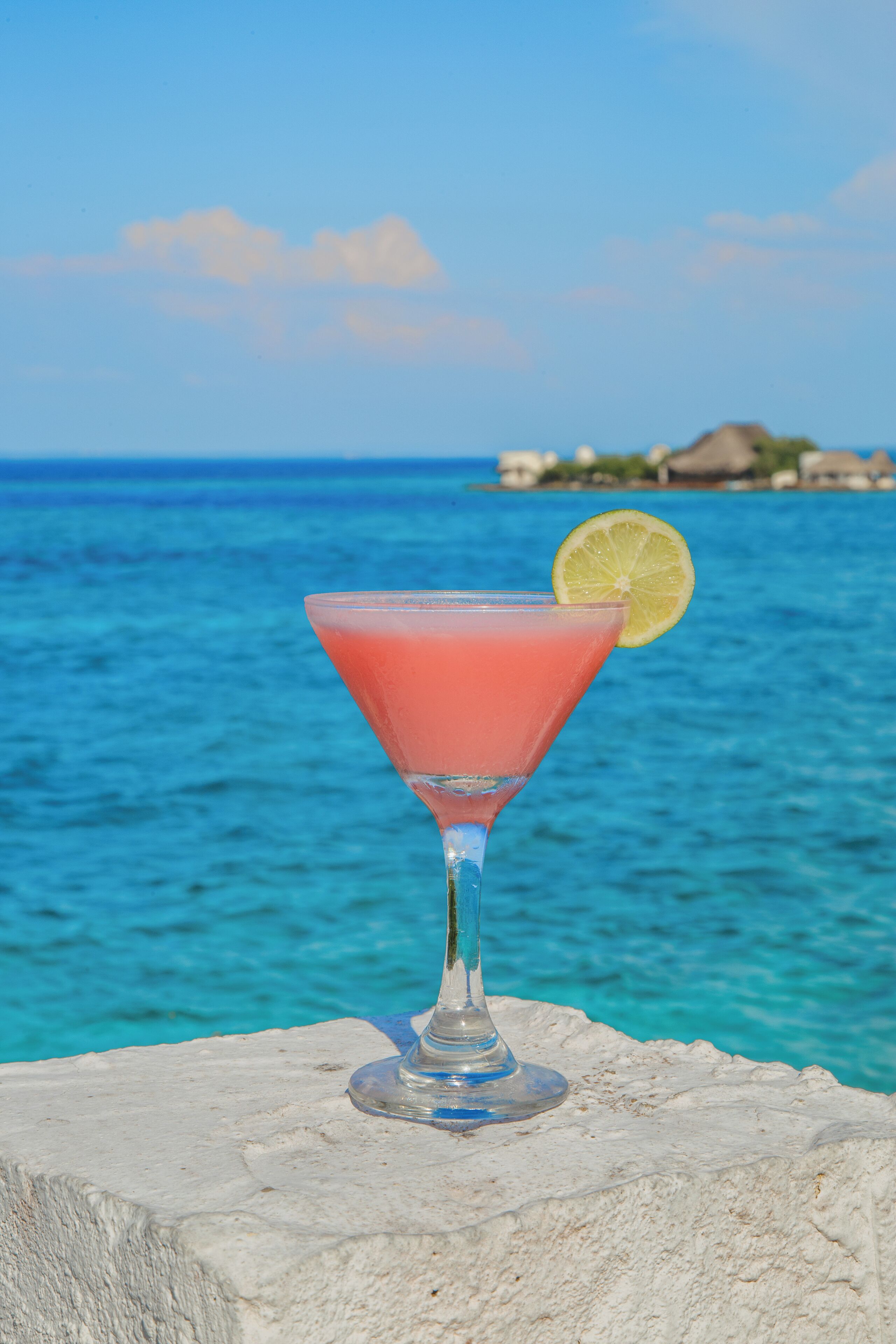 A cocktail glass with a slice of lime on a stone ledge by the sea.