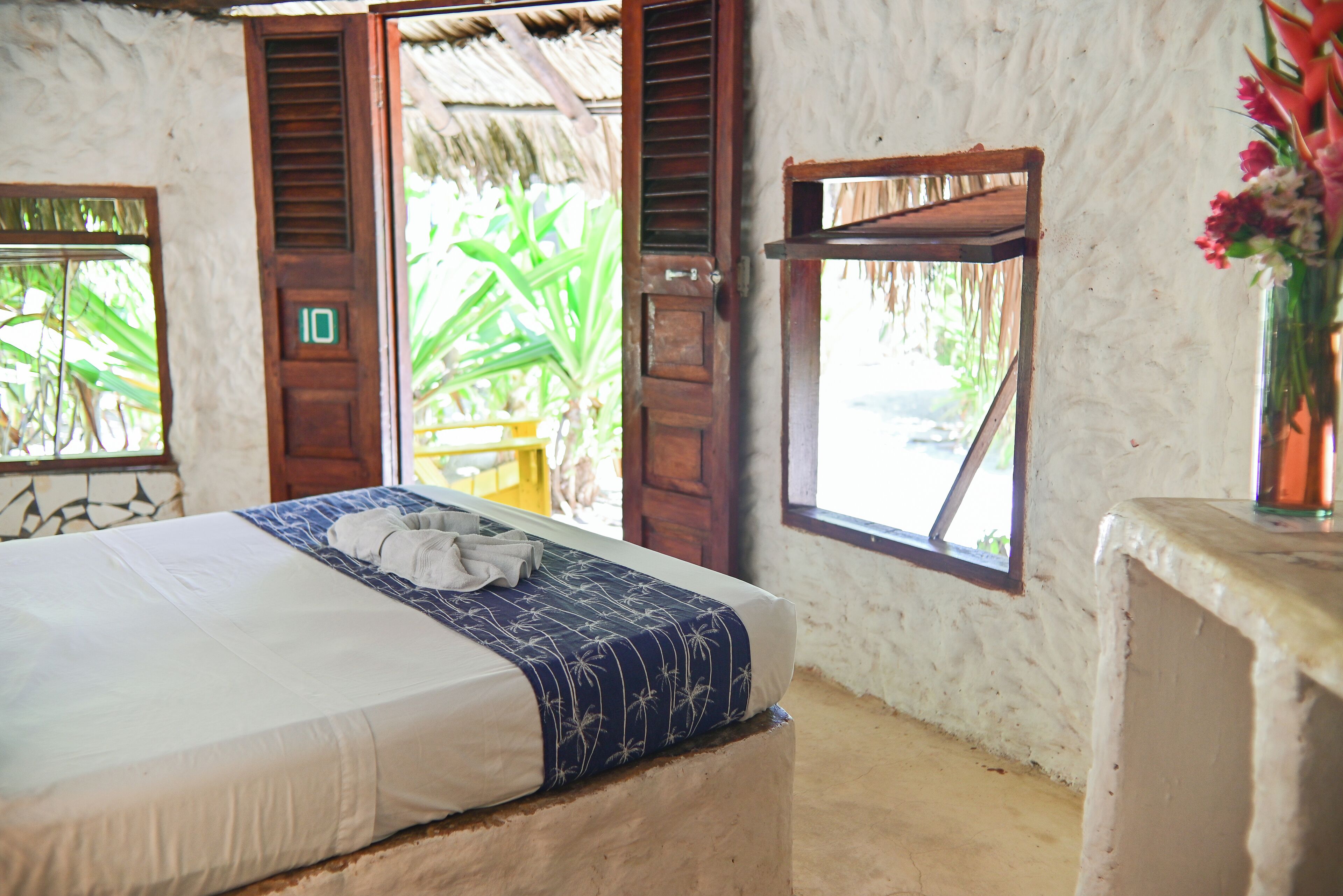 Um quarto com uma cama, uma porta de madeira com um número e uma janela com vista para o verde.