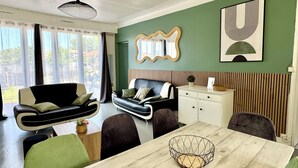 Ein modernes Wohnzimmer mit einer Couch, einem Esstisch und Stühlen. Es gibt ein großes Fenster mit Vorhängen, einen Spiegel an der Wand und ein gerahmtes Kunstwerk.