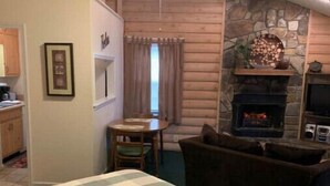 A bedroom with a bed, a ceiling fan, a stone fireplace, a dining table, and a view of the outdoors.