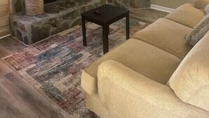 A living room with a stone fireplace, a flat-screen TV mounted on the wall, a beige sofa, and a patterned rug.