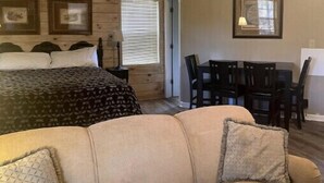 A cozy room with a bed, a beige sofa, a dining table with chairs, and wooden walls.