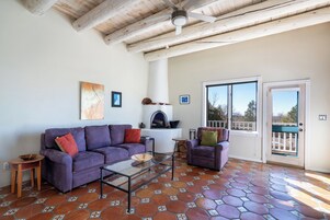 Un salón con un sofá morado, una mesa de centro de vidrio y una chimenea. Se ve una puerta con vistas al exterior.