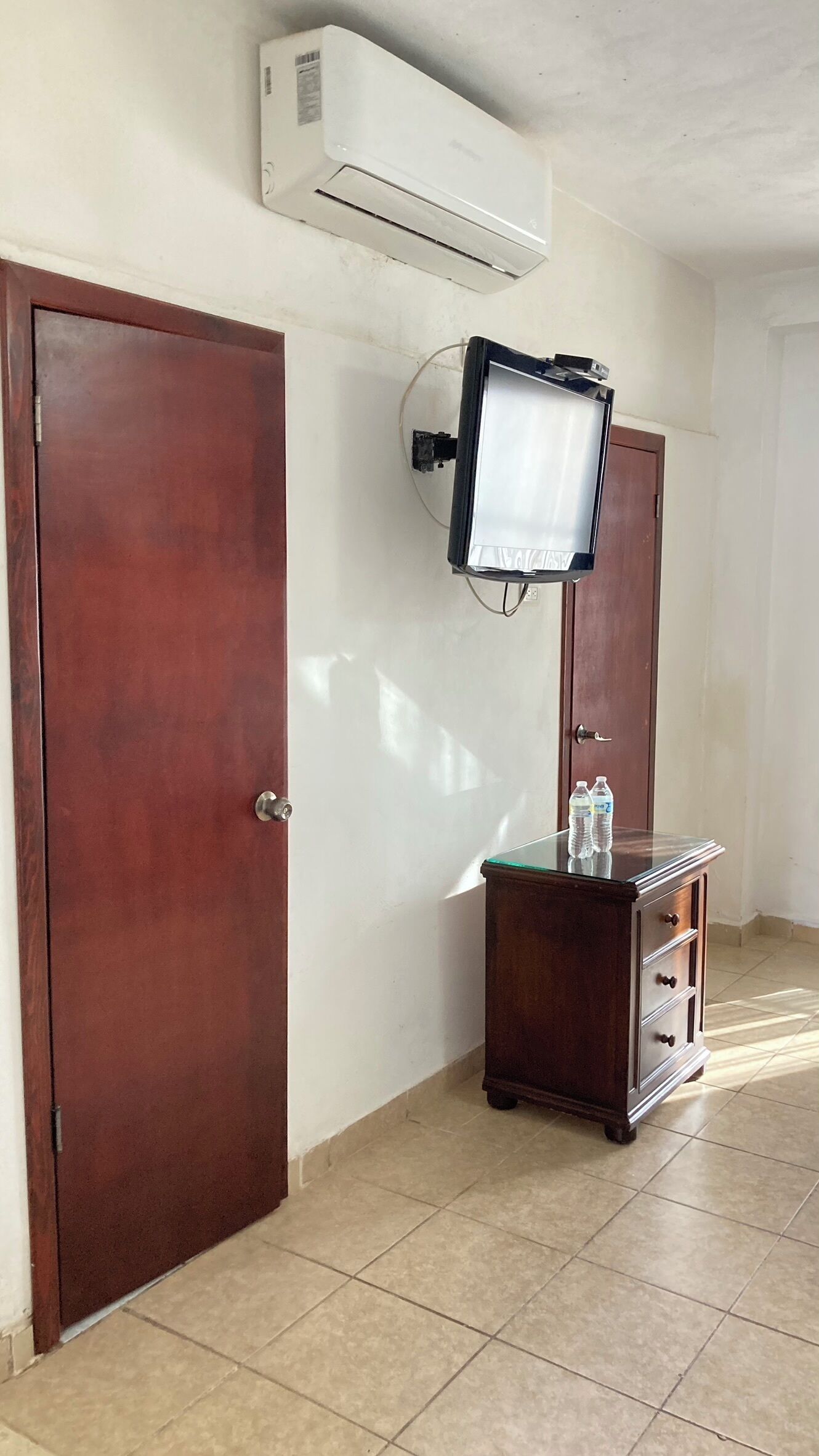 A hallway with two wooden doors, a small wooden cabinet with bottles, and an air conditioning unit mounted on the wall.