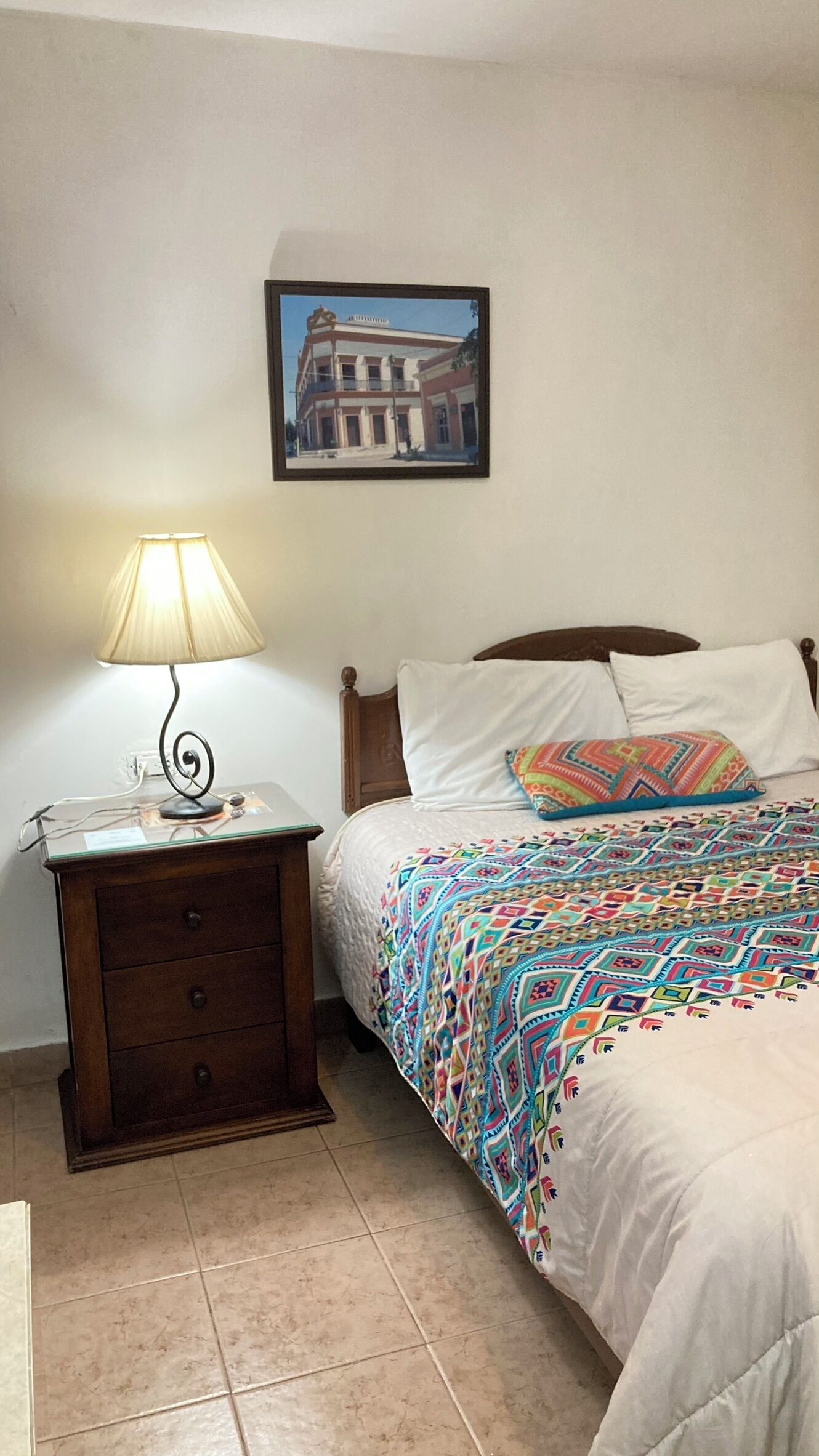 Ein Schlafzimmer mit einem Bett, einem Nachttisch mit Lampe, einer Kommode und einem gerahmten Bild an der Wand.