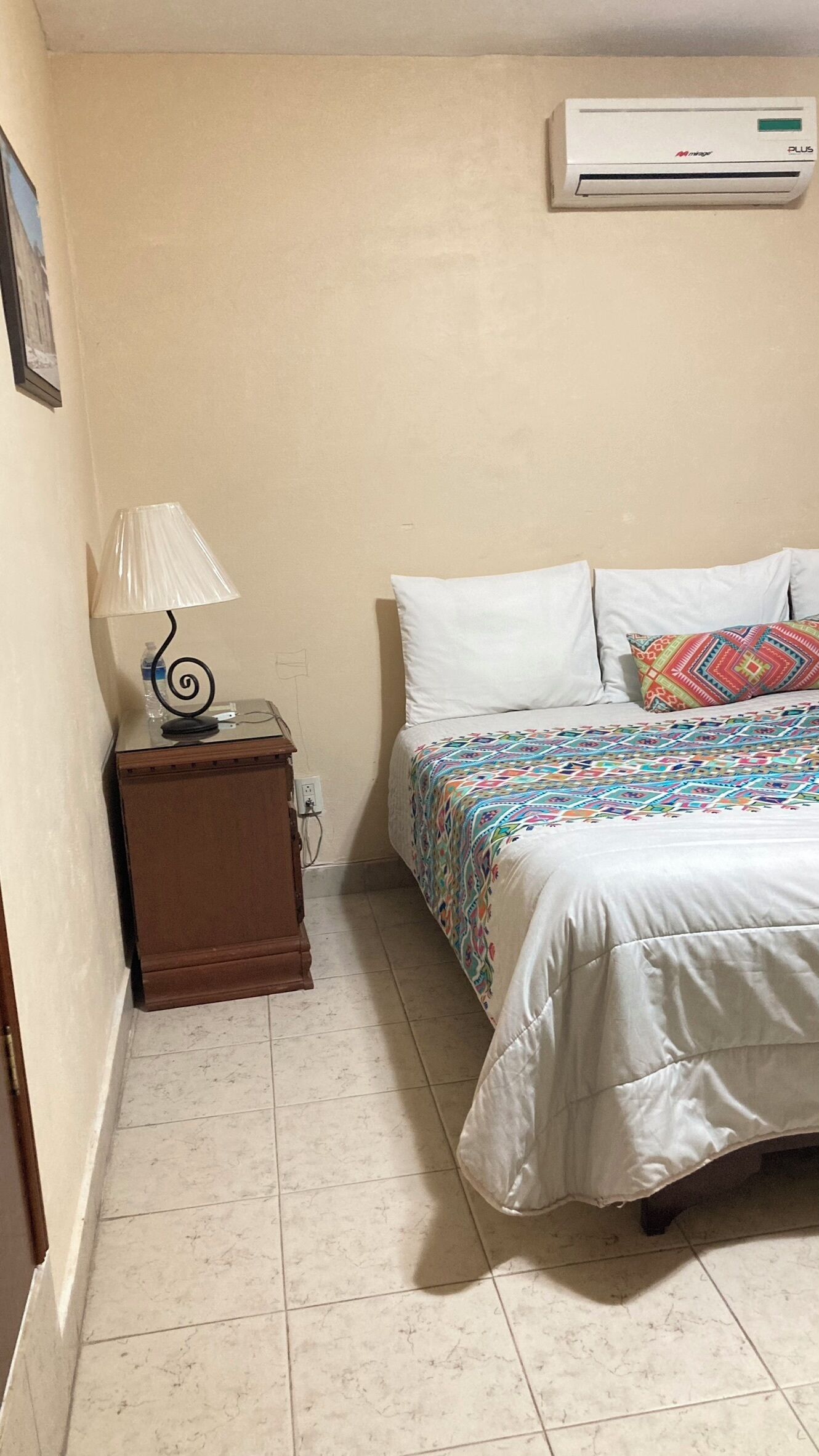 A bedroom with a bed, a nightstand, a lamp, and an air conditioner on the wall.