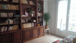 A room with a large wooden bookshelf filled with books and decorative items, a small white stool with a potted plant, and a large window providing natural light. - Cozy apartment in Paris (Paris)