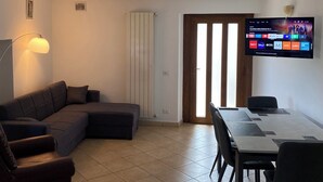 Un salon comprenant un canapé d’angle, une table à manger avec des chaises, un téléviseur à écran plat fixé au mur et une porte donnant sur une autre pièce.