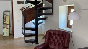 Un salon avec un fauteuil en cuir marron, un canapé gris, un escalier en colimaçon et une lampe blanche.