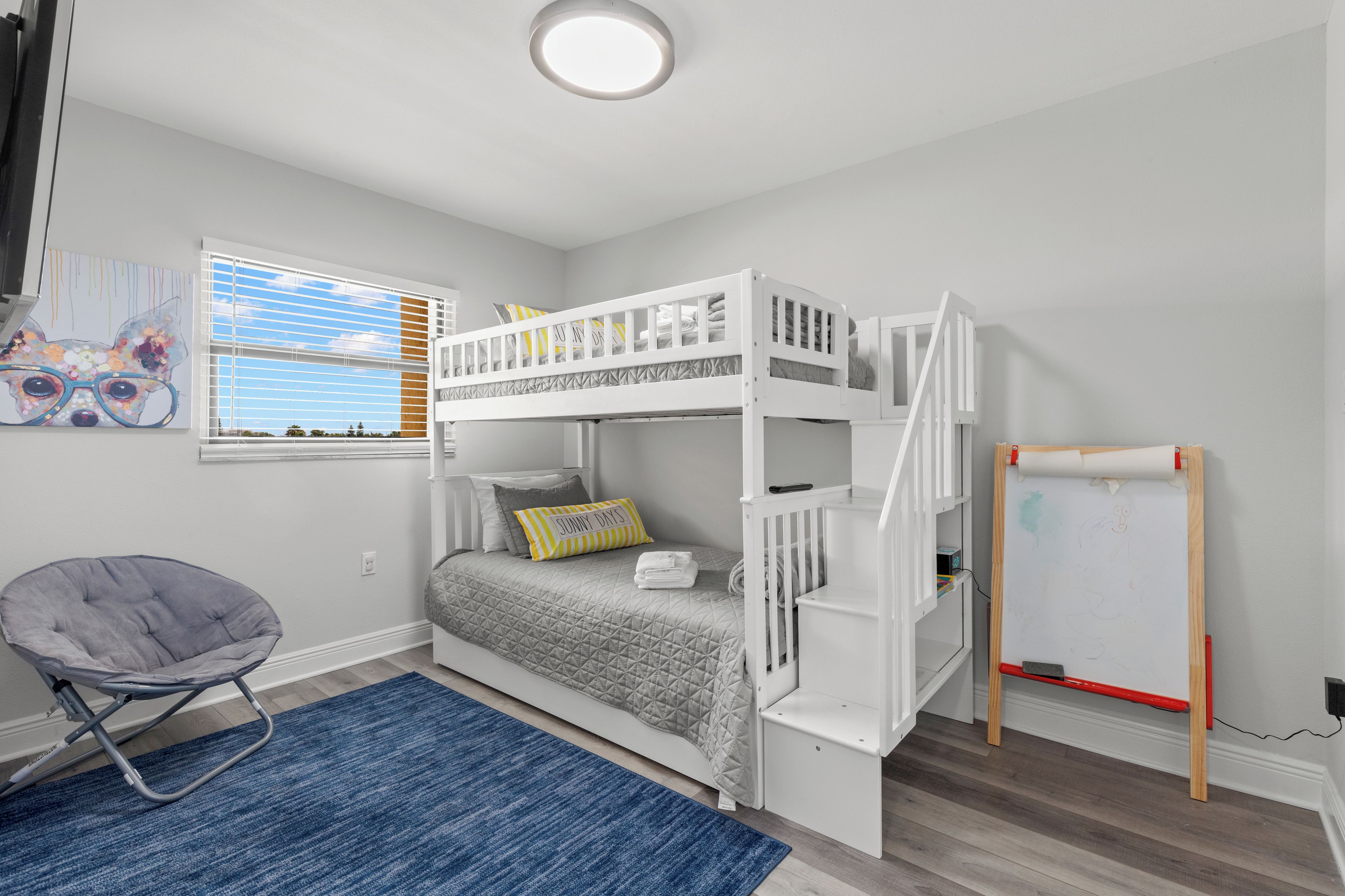 Une chambre avec un lit superposé, un bureau, une chaise, un tableau blanc et une fenêtre avec des stores.