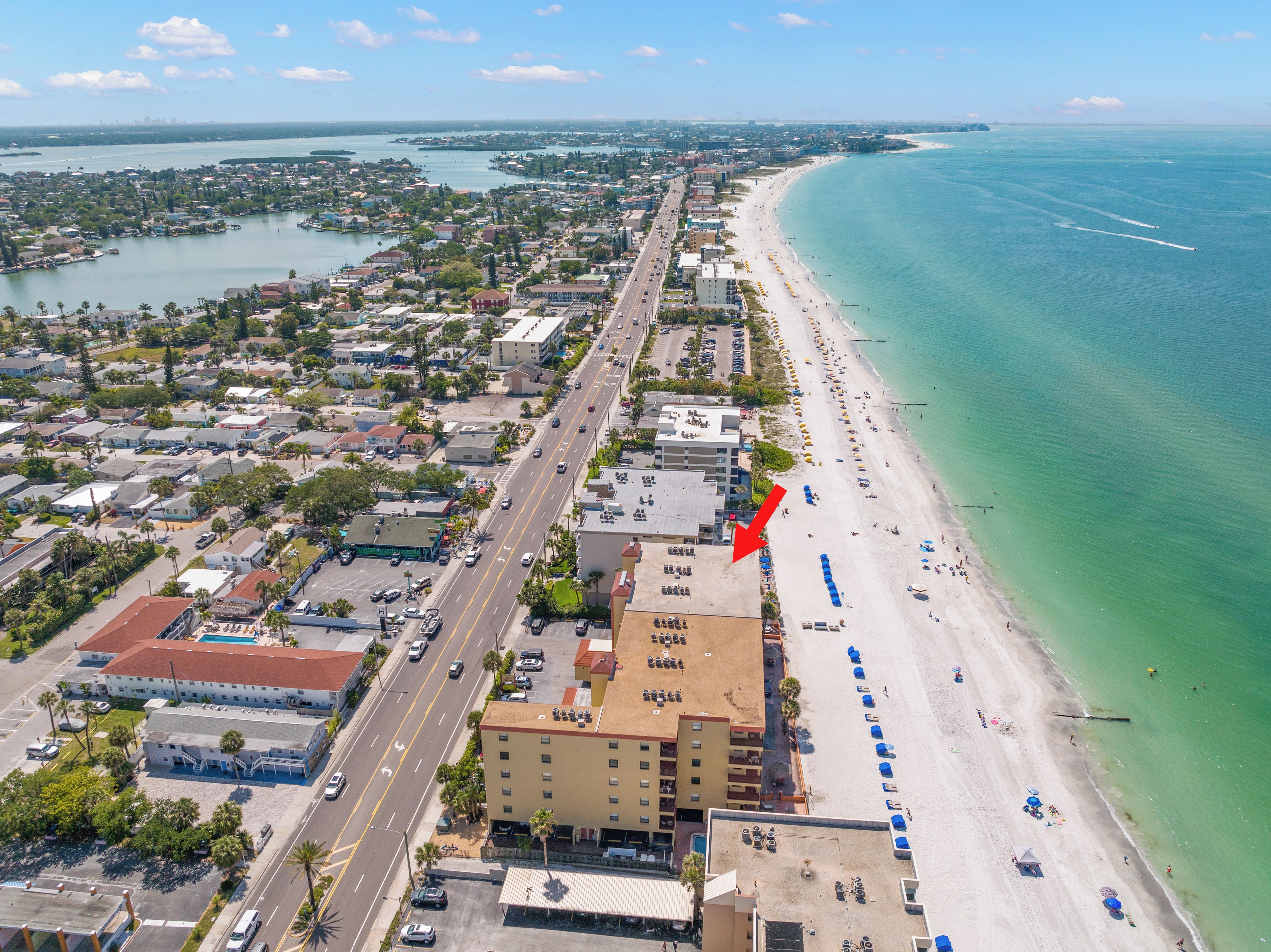 An aerial view of a coastal city with a long beach, a prominent road running parallel to the shore, and a hotel with a pool area.