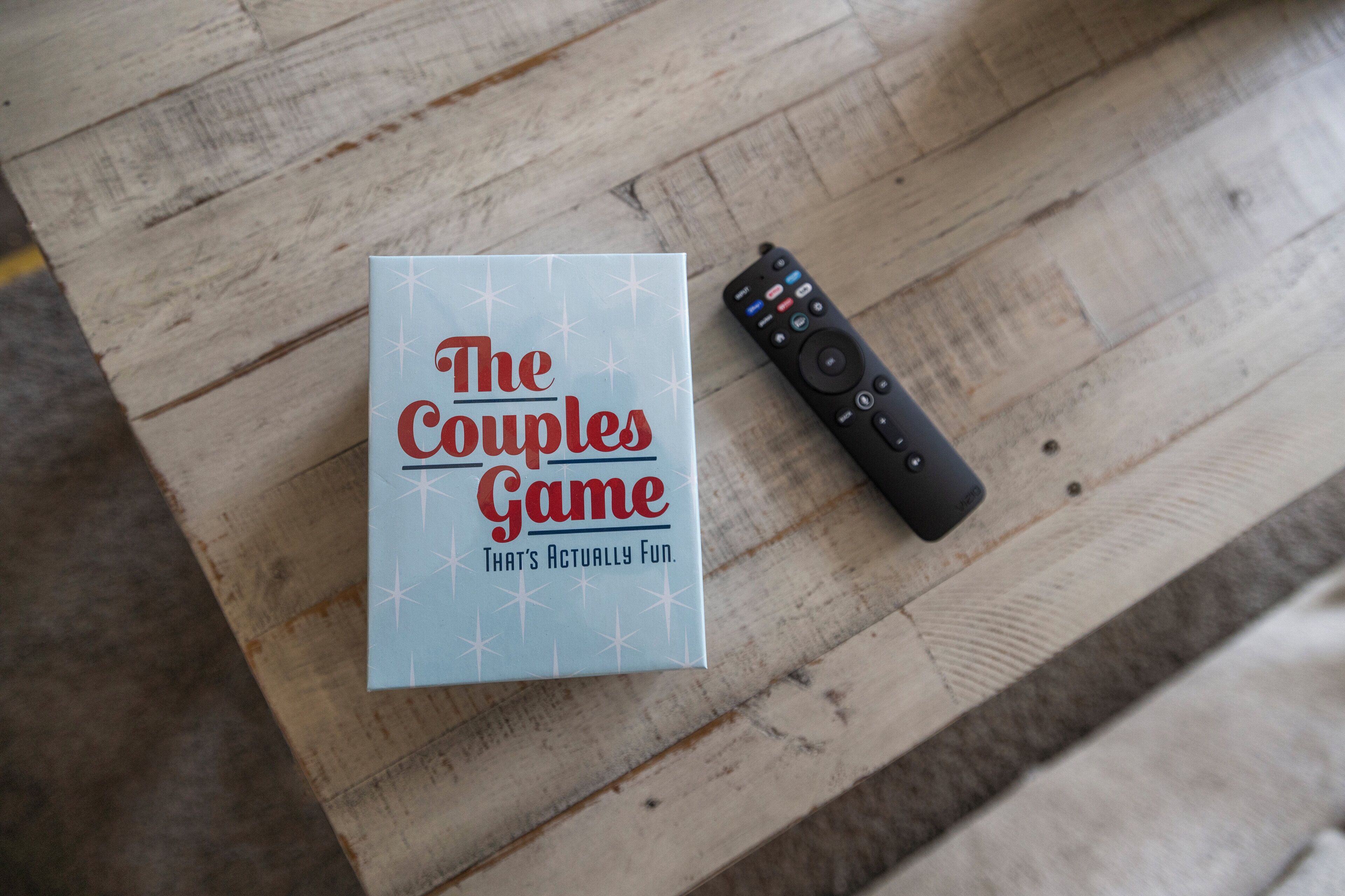 Een speldoos met de titel 'The Couples Game' en een afstandsbediening op een houten ondergrond.