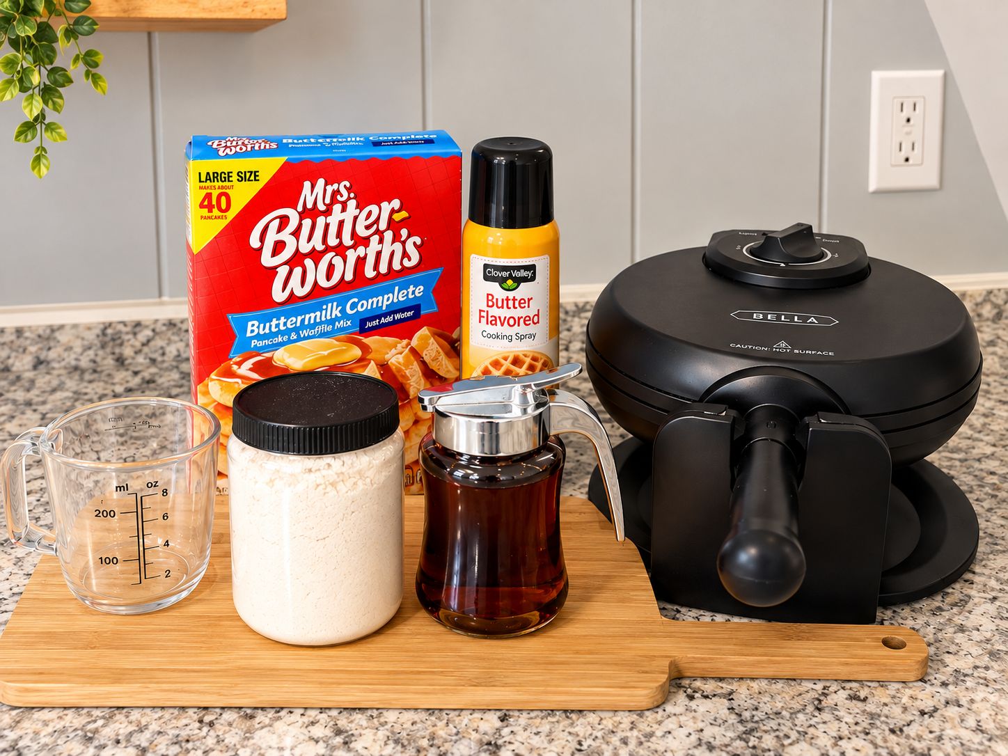 Una encimera de cocina con una caja de mezcla para panqueques y waffles Mrs. Butterworths Buttermilk, un aerosol de sabor a mantequilla, un vaso medidor, una botella de suero de leche y una plancha para waffles.