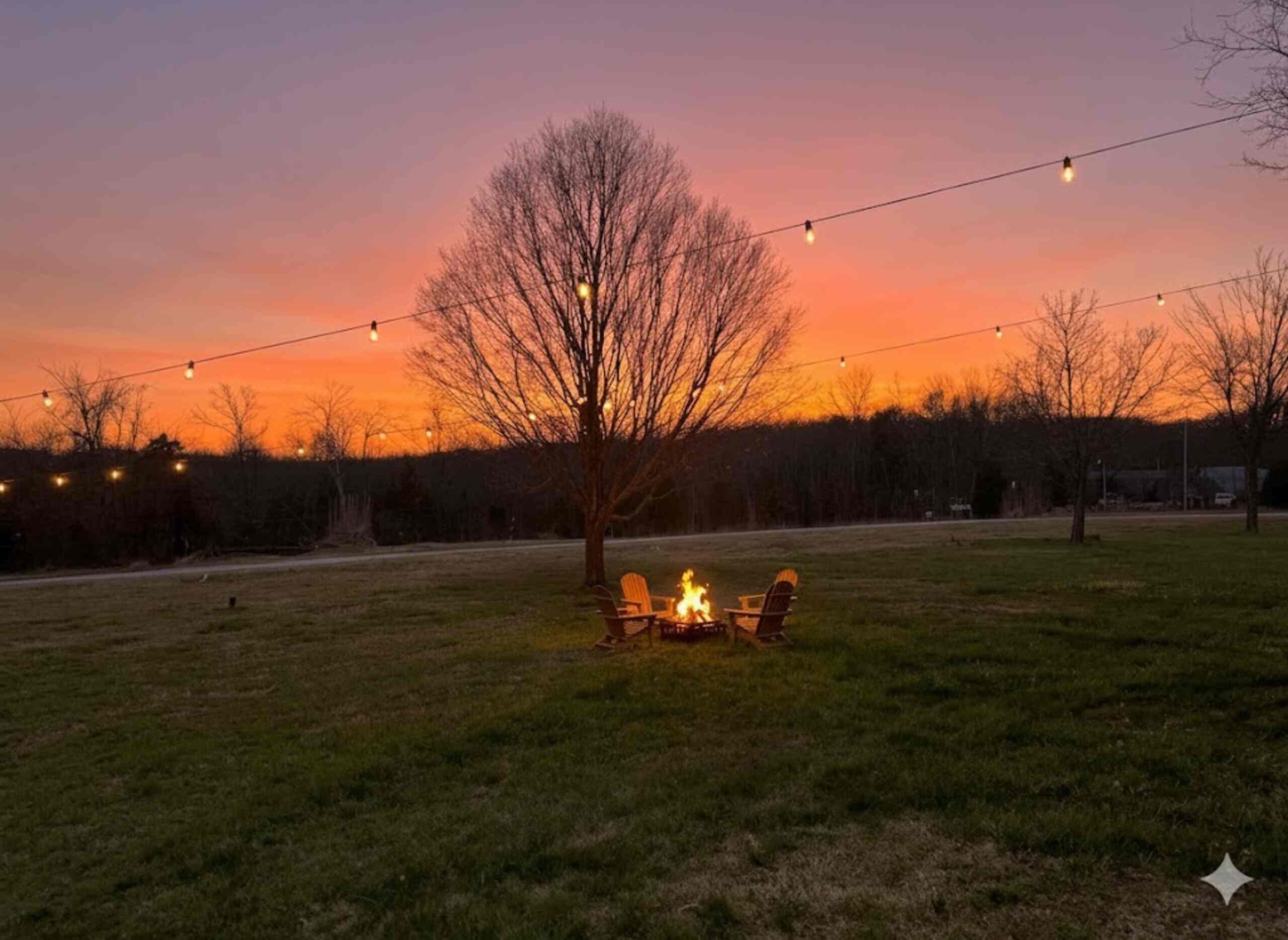An outdoor setting with a fire pit and chairs, surrounded by a grassy area and trees, under a twilight sky.