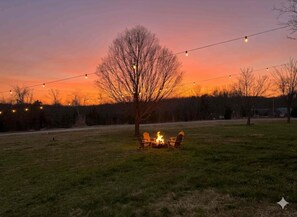 An outdoor setting with a fire pit and chairs, surrounded by a grassy area and trees, under a twilight sky. - A-frame for 2, Waffle Bar, Fire pit, Walk 2 Baxter Marina (Lampe)