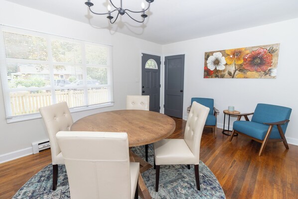 A dining area with a round wooden table, four white chairs, a blue rug, and a floral painting on the wall.