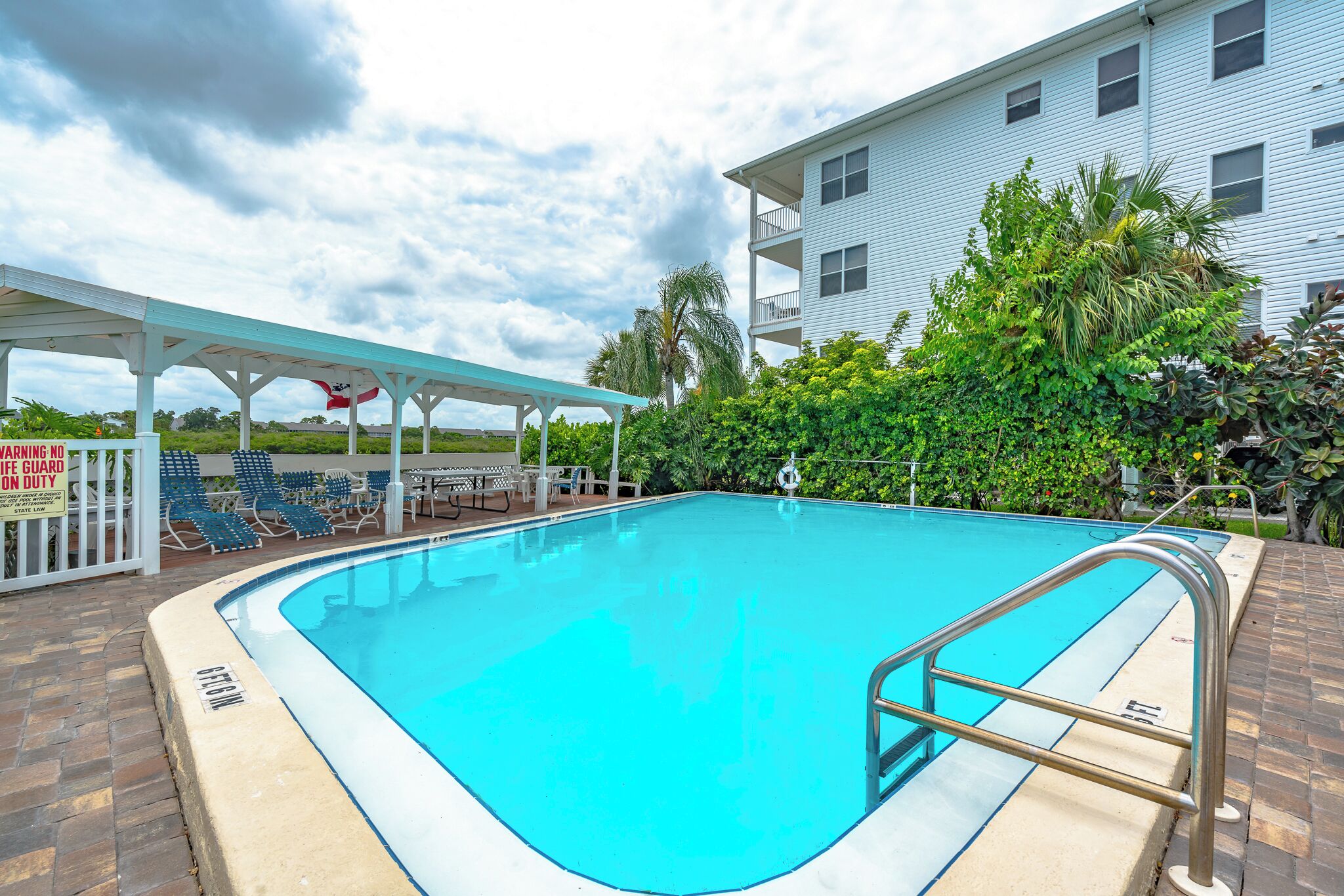 A swimming pool with a white deck, a covered seating area, and a building with balconies.