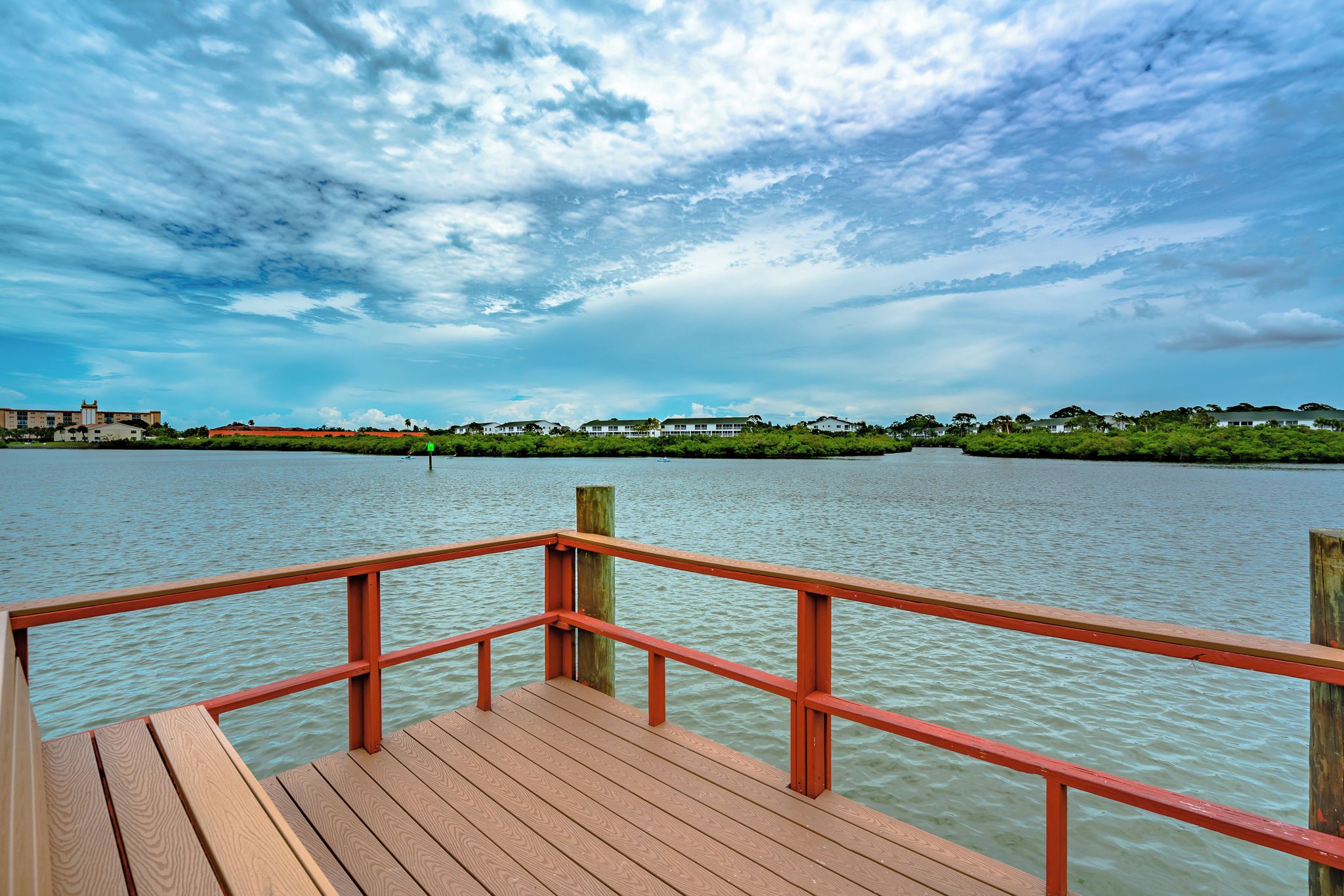 A wooden pier with a red railing extending over calm water, leading to a shoreline with buildings and greenery.