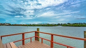 Un muelle de madera con barandilla roja se extiende sobre aguas tranquilas, llegando a la orilla con edificios y vegetación.