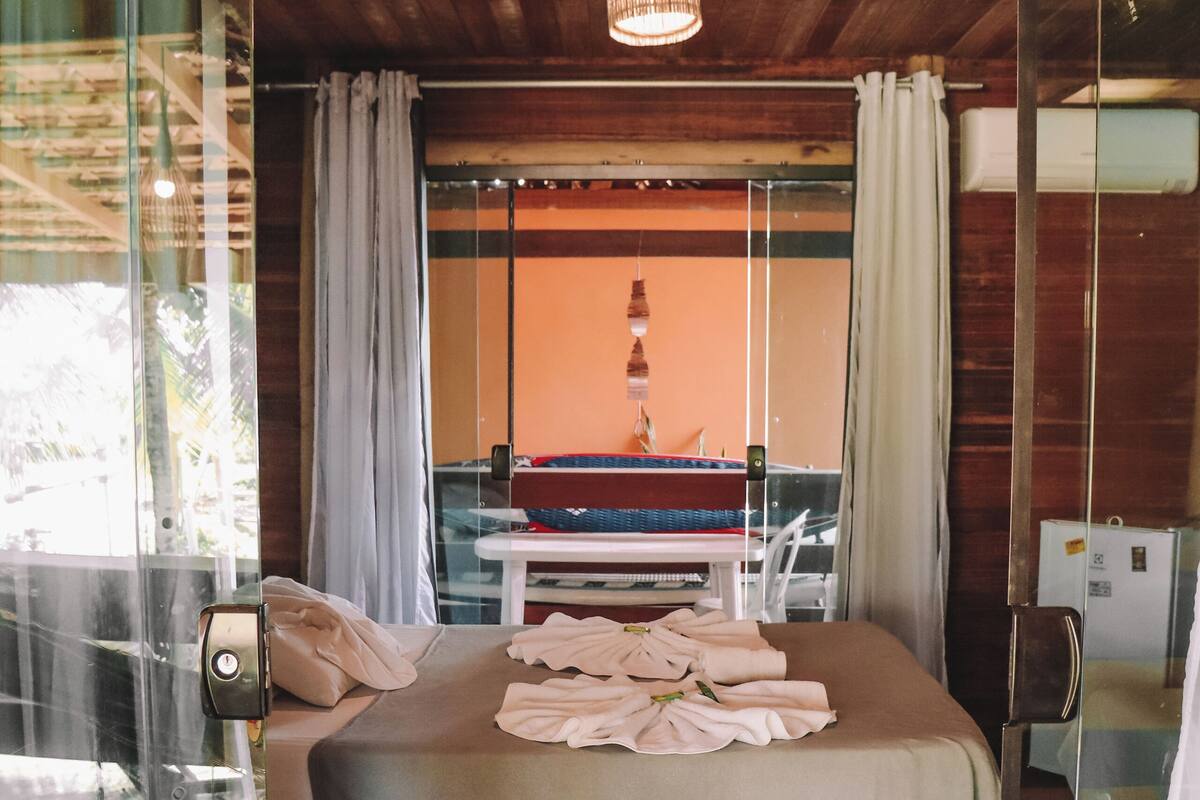 Une chambre avec un plafond en bois, un lit recouvert d’une courtepointe blanche, une table de chevet et une vue sur une pièce où se trouvent une chaise rouge et une table.