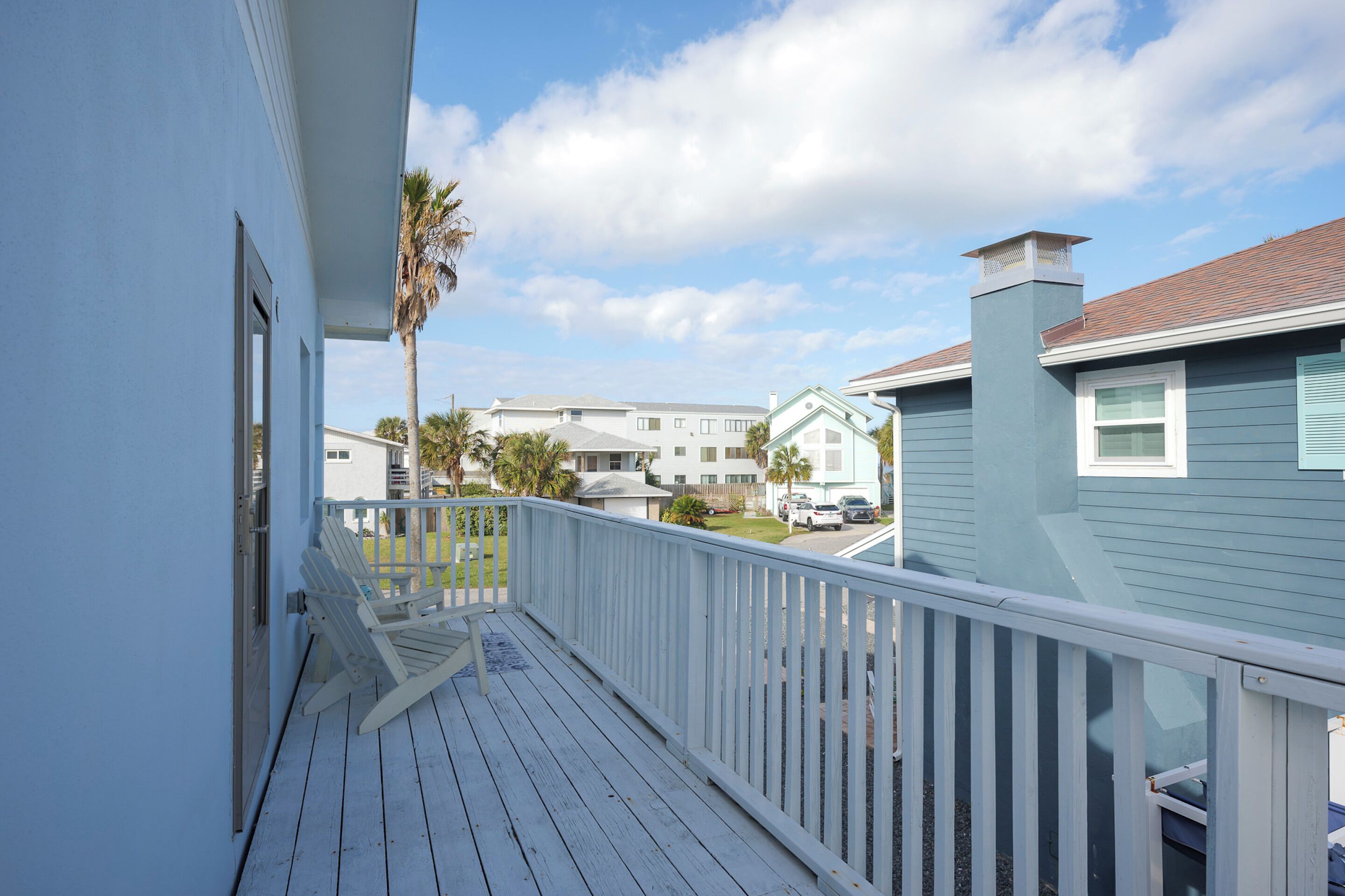 Um deck de madeira com guarda-corpo branco, com vista para uma área residencial com casas e céu claro.