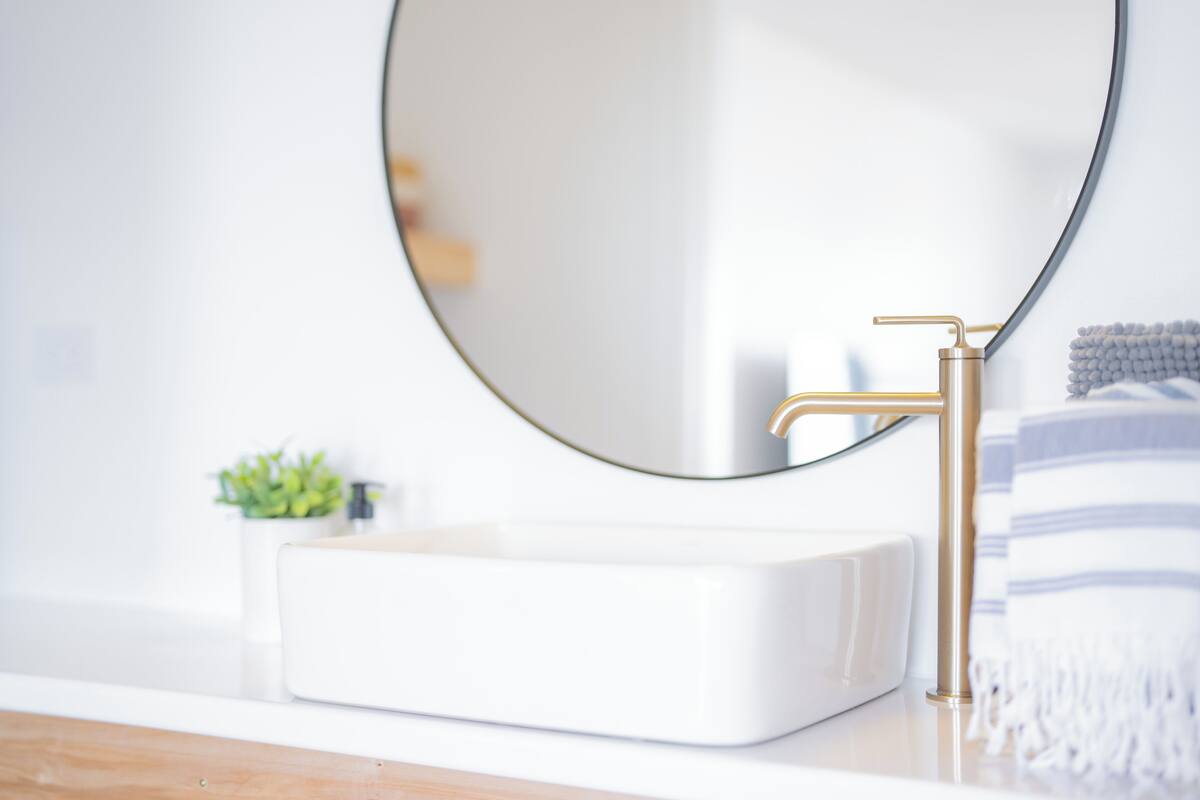 A bathroom with a white sink, gold faucet, and a round mirror.