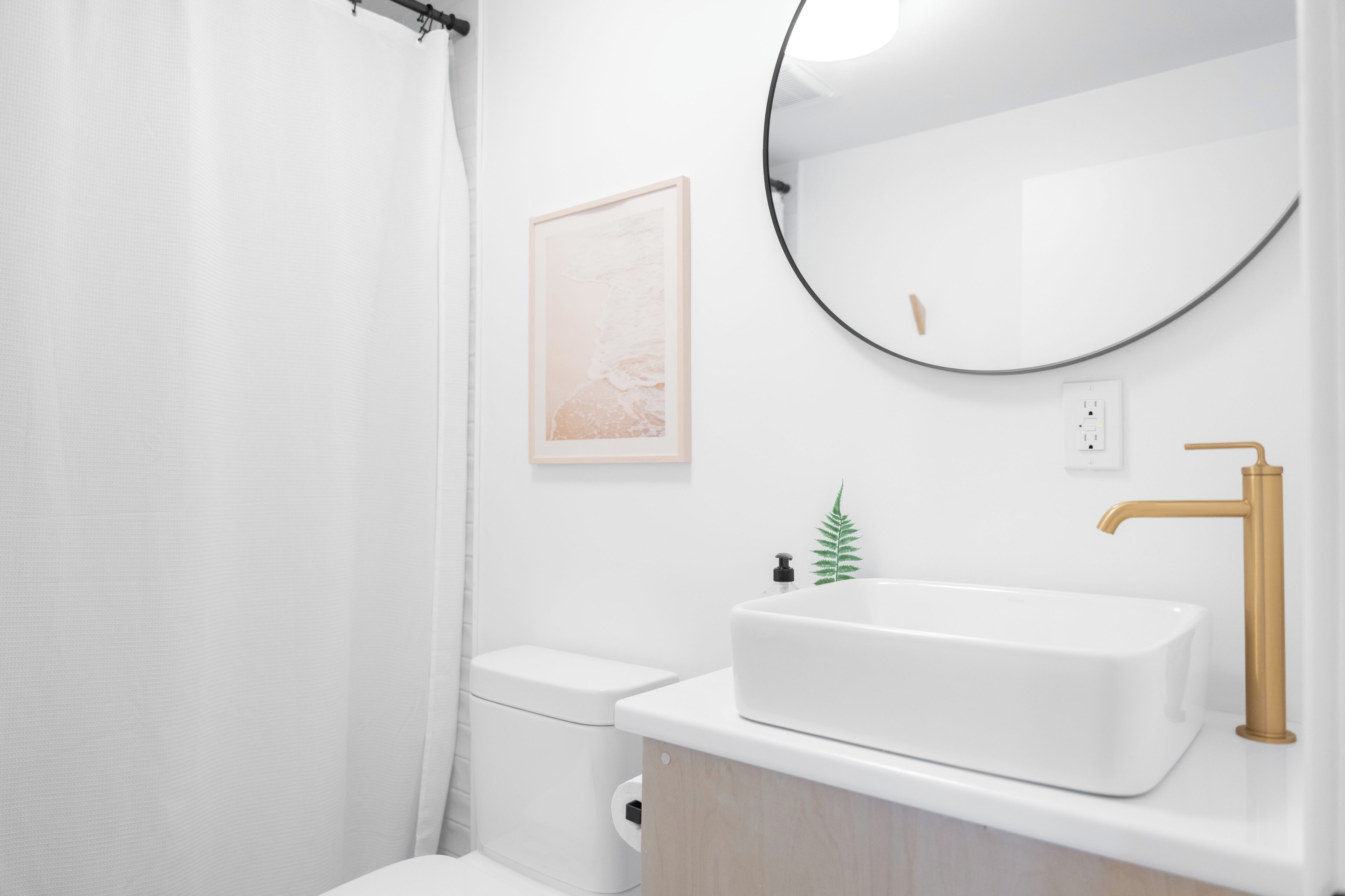 A modern bathroom with a white sink, gold faucet, and a round mirror.