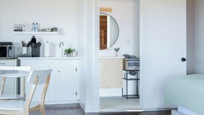 Cozinha moderna com armários brancos, micro-ondas e máquina de café. Possui uma área de jantar com mesa e cadeiras brancas. É visível um quarto com cama e porta de acesso a um banheiro.