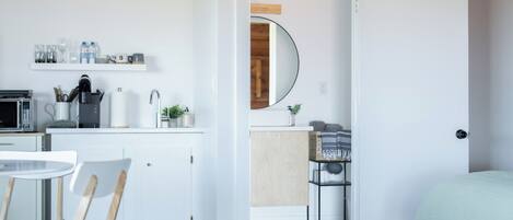 Une cuisine moderne avec des meubles blancs, un four à micro-ondes et une machine à café. On y trouve un coin repas avec une table et des chaises blanches. Une chambre à coucher avec un lit et une porte donnant sur une salle de bain est visible.