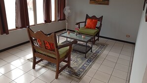 Una sala de estar con un sofá, una silla, una mesa de centro de vidrio, un ventilador y un cuadro en la pared.