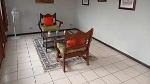 Una habitación con suelo de baldosas, dos sillas de madera con cojines, una mesa de vidrio, un ventilador de pie y tres cuadros en la pared.